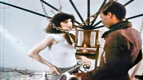 Gagged skinny brunette in white tank top bound by chains as man in leather holds whip in retro BDSM scene