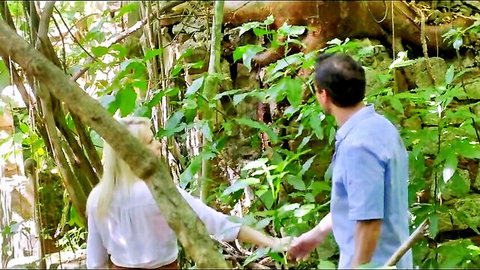 Blonde babe in white blouse holds hands with handsome guy in lush jungle outdoor romance scene