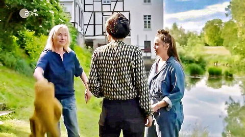Clothed blonde woman holding teddy bear walks with man and brunette by riverside house