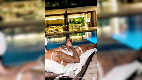 Tattooed olive-skinned Latina with medium tits lies by pool spreading legs fingering pussy in white thong