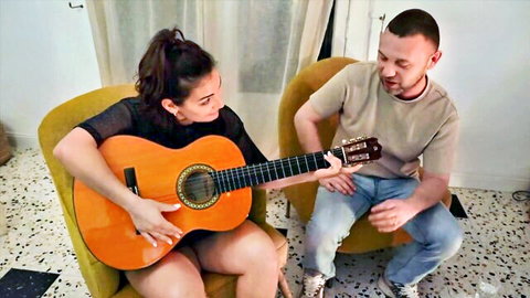 Brunette in sheer black top and mini dress strums orange guitar seductively beside smiling man on chairs
