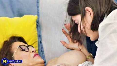 Young brunette in glasses squeezes mature woman's huge exposed tits on bed
