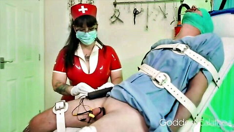 Red latex nurse Goddess Kaline holds electro device on bound patient's exposed cock with electrodes in femdom medical play