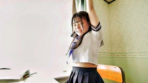 Smiling Asian schoolgirl in sexy sailor uniform raises arms teasing in classroom