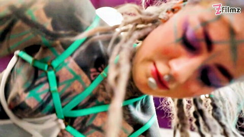 Tattooed dreadlock woman in green harness exposing inked tits and body with intense makeup