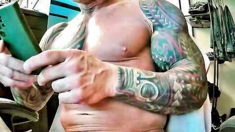 Tattooed muscular man holding phone shirtless in gay solo tease