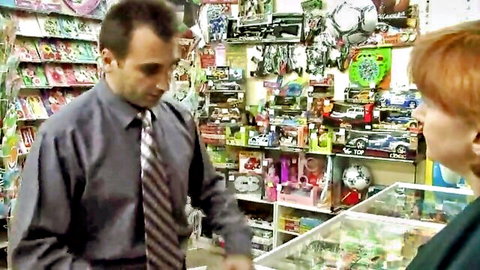 Suited man leans in to talk with smiling redhead woman at toy store counter