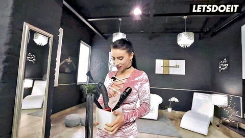Brunette in revealing pink lace dress holding two big black dildos in white vase smiling seductively