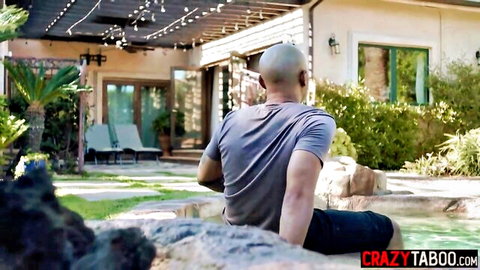 Muscular bald man in gray shirt sitting poolside by outdoor spa at night