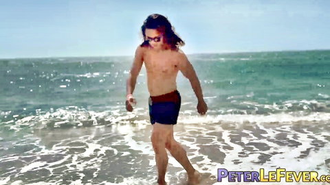 Shirtless long-haired young hunk walks on beach in tight shorts showing muscular body