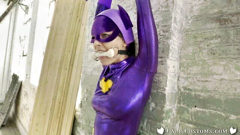 Gagged purple superheroine in shiny latex catsuit with arms raised against wall teasing cosplay