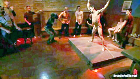 Muscular naked man poses with hard cock arms raised on stage surrounded by watching men in gay club