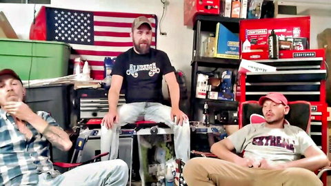 Three bearded men in casual clothes hanging out in a cluttered garage workshop with tools and American flag.