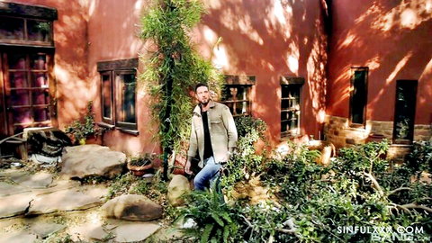 Handsome man in beige jacket and jeans posing confidently outdoors by red adobe building with plants