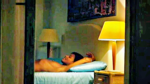 Nude ebony woman lying topless on bed with perky tits exposed in dim bedroom glow