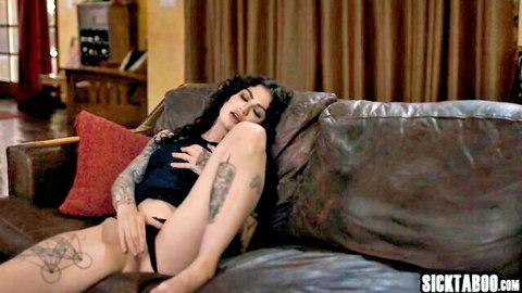 Tattooed brunette lies on couch spreading legs wide exposing shaved pussy in pulled-aside black thong