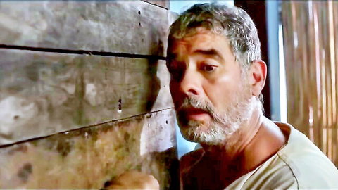Gray-haired mature man with beard leans against rustic wooden wall peeking intensely