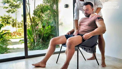 Older daddy gropes muscular young guy's pecs and shoulders as he sits spread-legged in tight shorts