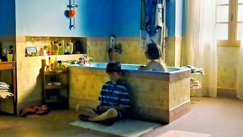 Topless woman relaxes in bubbly bathtub while young boy sits watching nearby in steamy bathroom