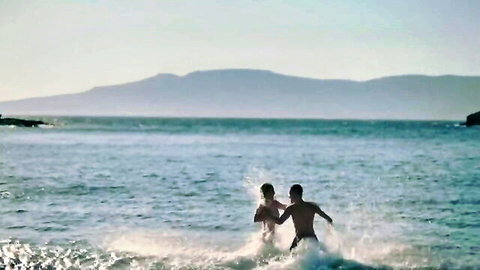Topless young couple splashing playfully in shallow turquoise ocean waves at sunset
