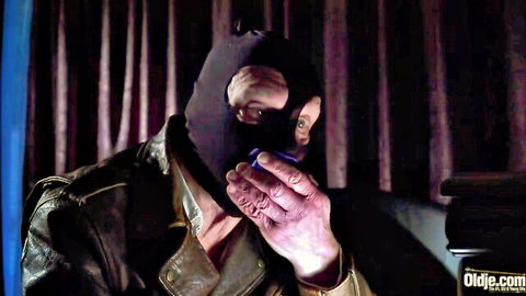 Masked old man in leather jacket holds cigarette to mouth in dim room