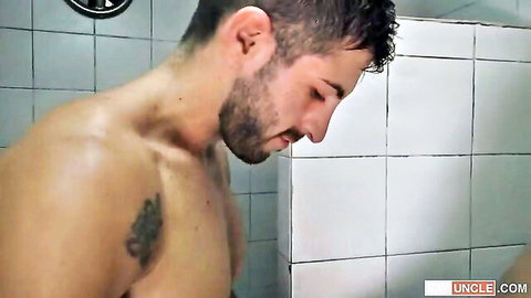 Muscular bearded man with tattoo showering shirtless in tiled bathroom