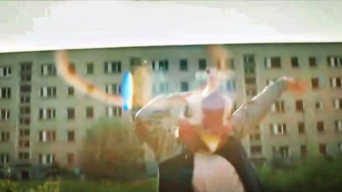 Tattooed man posing arms outstretched in front of gray apartment building with colorful lens flares