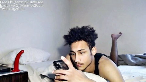 Young muscular black guy topless on bed holding phone next to red dildo