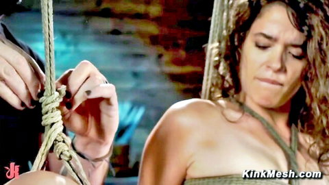 Topless woman with exposed tits hogtied in suspension rope bondage by hands in dungeon