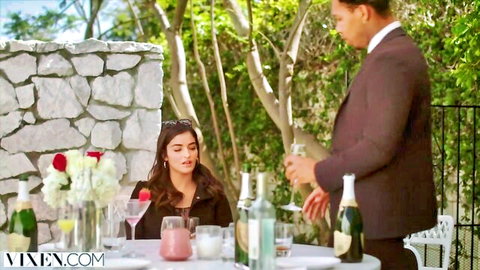 Brunette woman in sunglasses toasts champagne with suited man at outdoor patio table