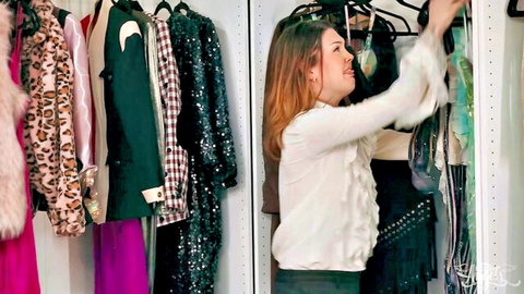 Brunette babe in ruffled white blouse reaches up grabbing sparkly dresses in crowded closet