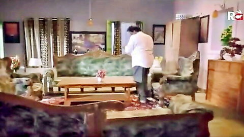Clothed South Asian man stands with hands clasped in ornate Indian living room with green velvet sofas and wooden table.