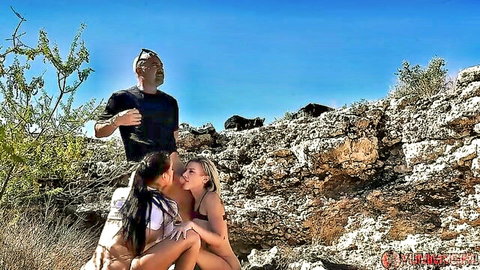 Busty MILFs in bikinis kissing passionately in hot desert with watching man