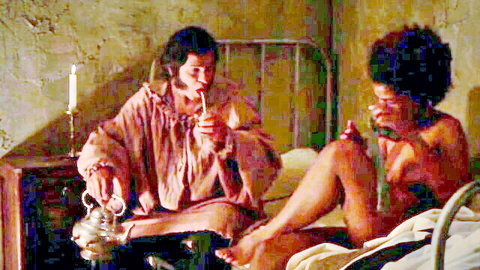 Nude ebony woman with afro spreads legs topless on bed while robed brunette smokes pipe in vintage room