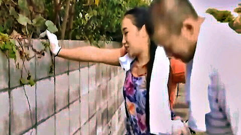 Smiling Asian woman in floral top and gloves reaches for vines outdoors with man in towel