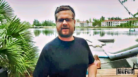 Bearded man in glasses and cap smiles outdoors by tropical lake dock with boats