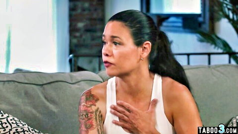Tattooed brunette MILF in tight white tank top looks shocked with hand on chest in taboo scene