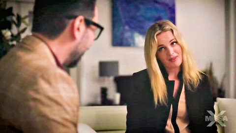 Blonde MILF in black suit smiling seductively at man in brown jacket on couch