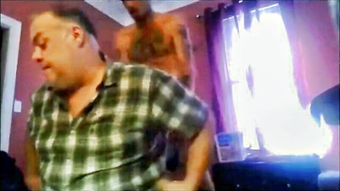 Chubby gay dude in open plaid shirt grins as tattooed muscular hunk stands close behind him