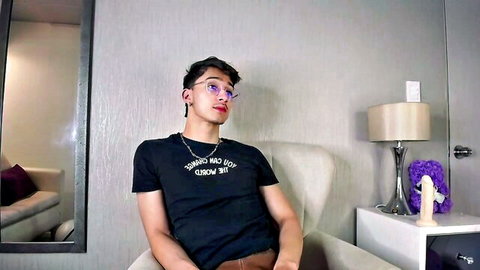 Sexy Latino guy in glasses and bad bitch shirt sits teasingly with big dildo on table in webcam show