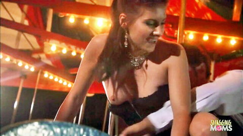 Busty brunette in strapless black dress flashes deep cleavage at outdoor carnival with man behind her