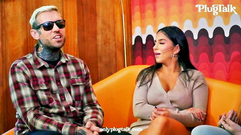 Tattooed blond guy in sunglasses chats with busty brunette babe on couch