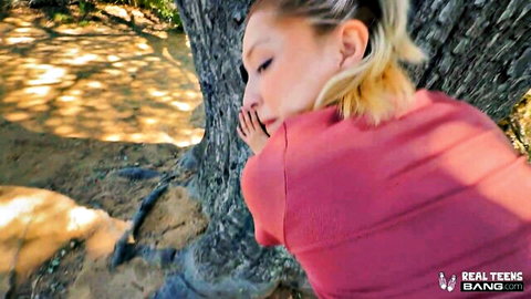 Blonde girl in red top leans seductively against tree outdoors, POV tease from hot threesome