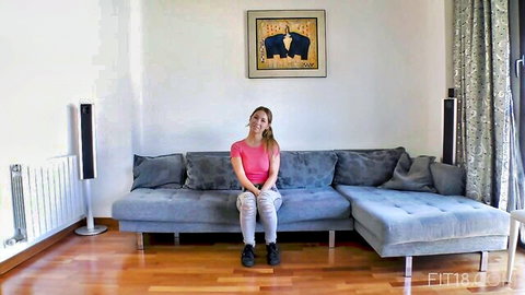 Smiling Caucasian brunette in tight pink top and khaki pants sitting on gray couch teasing