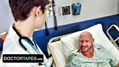 Male doctor in white coat with stethoscope examines bald muscular patient in hospital gown on bed