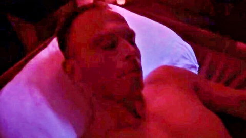 Shirtless bald man lying relaxed on pillows in neon red purple lit bedroom