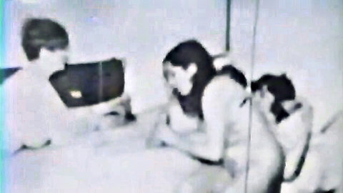Vintage black and white nude brunette with big tits smiles at naked shaggy haired guy while another nude woman kneels behind
