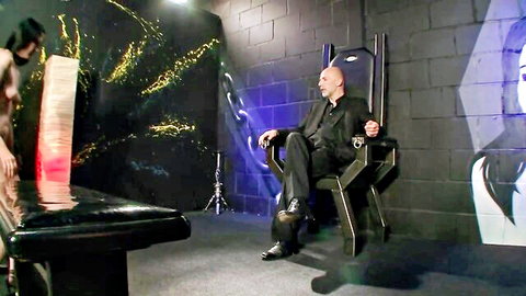 Bald suited dom holds leashes of two topless big-titted hooded slaves kneeling submissively in BDSM throne room
