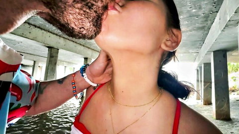 Bearded man grips and passionately kisses sexy young brunette's neck under pier