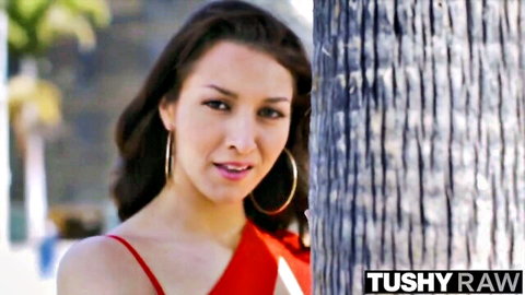 Brunette beauty in red dress smiles by palm tree in TUSHY RAW anal threesome teaser